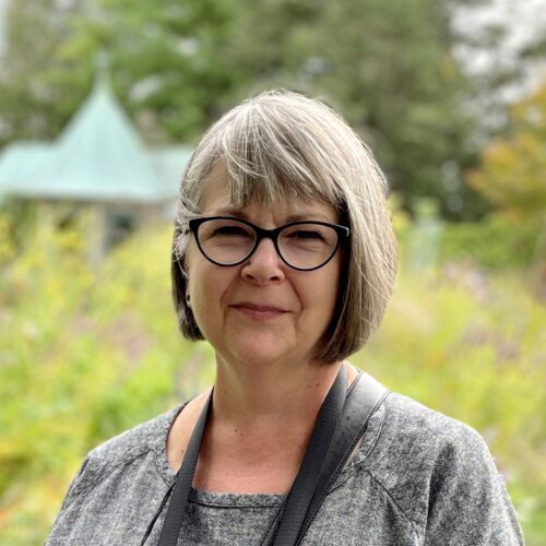 A woman with short gray hair and bangs is wearing glasses and a black camera strap around her neck. She is looking right at the camera with a slight smile.